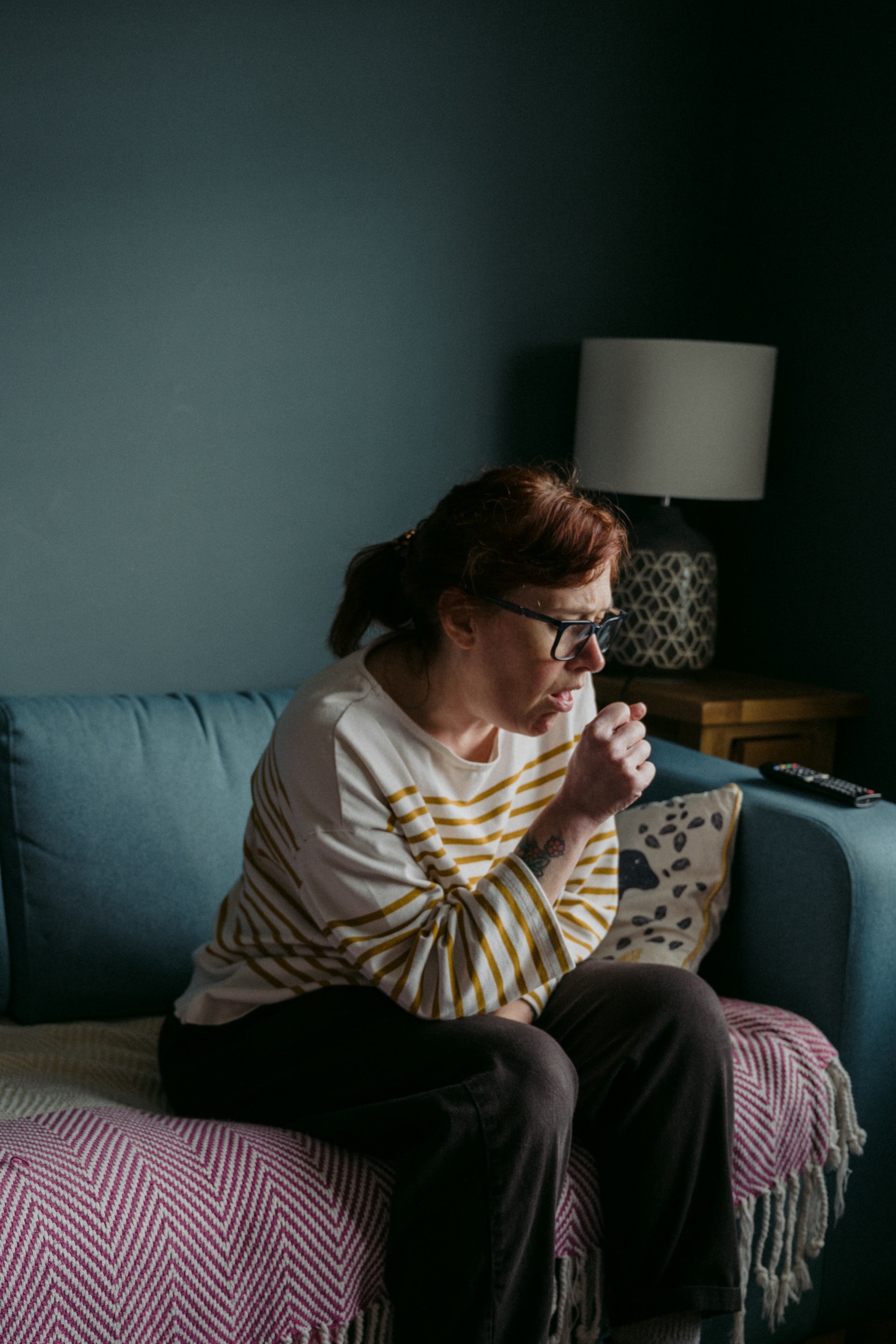 A woman with auburn hair sits on a sofa coughing