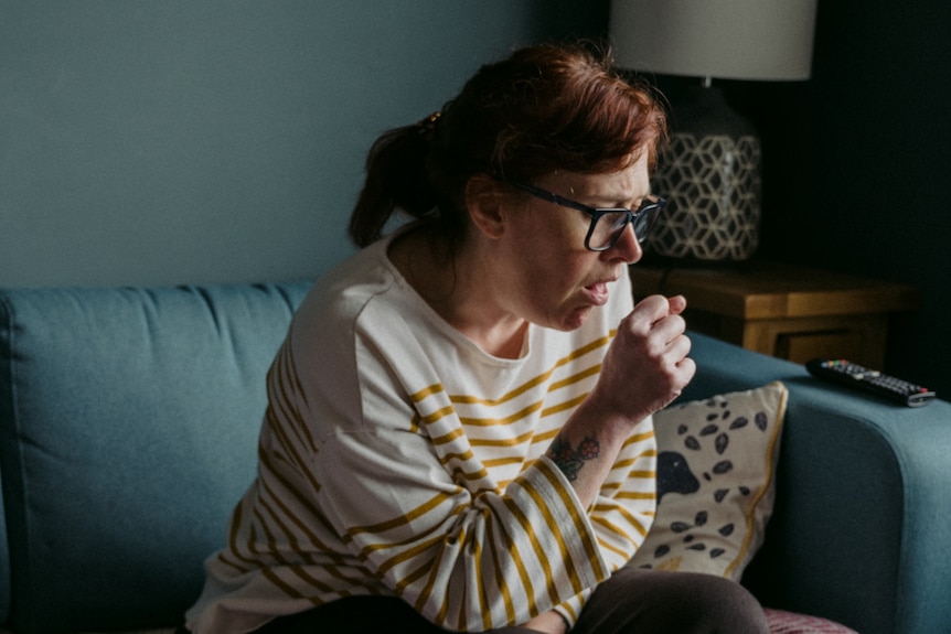 A woman with auburn hair sits on a sofa coughing