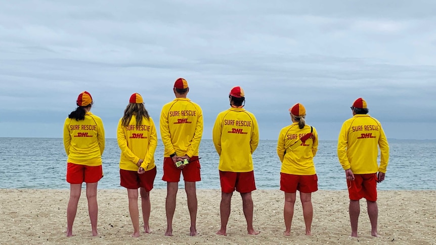 Six lifesavers standing in a row looking out to sea.