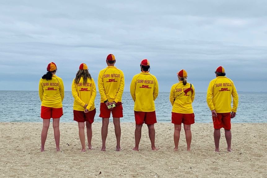 Six lifesavers standing in a row, looking out to sea.