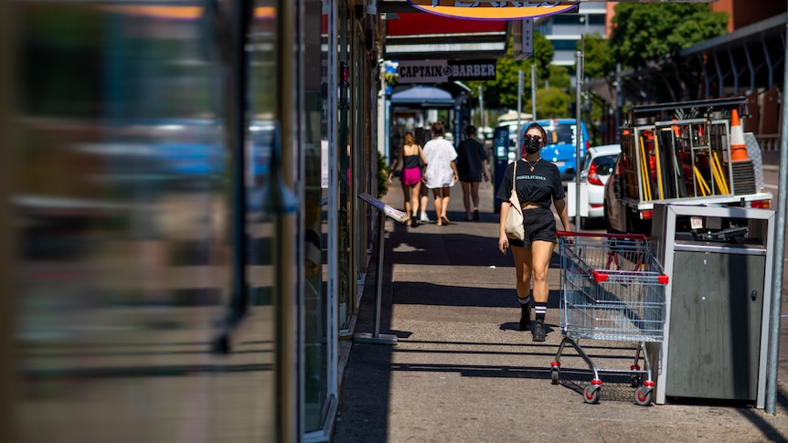 A woman wears a face mask while walking in Darwin CBD. She is wearing black shorts and a black top. It is a sunny day.