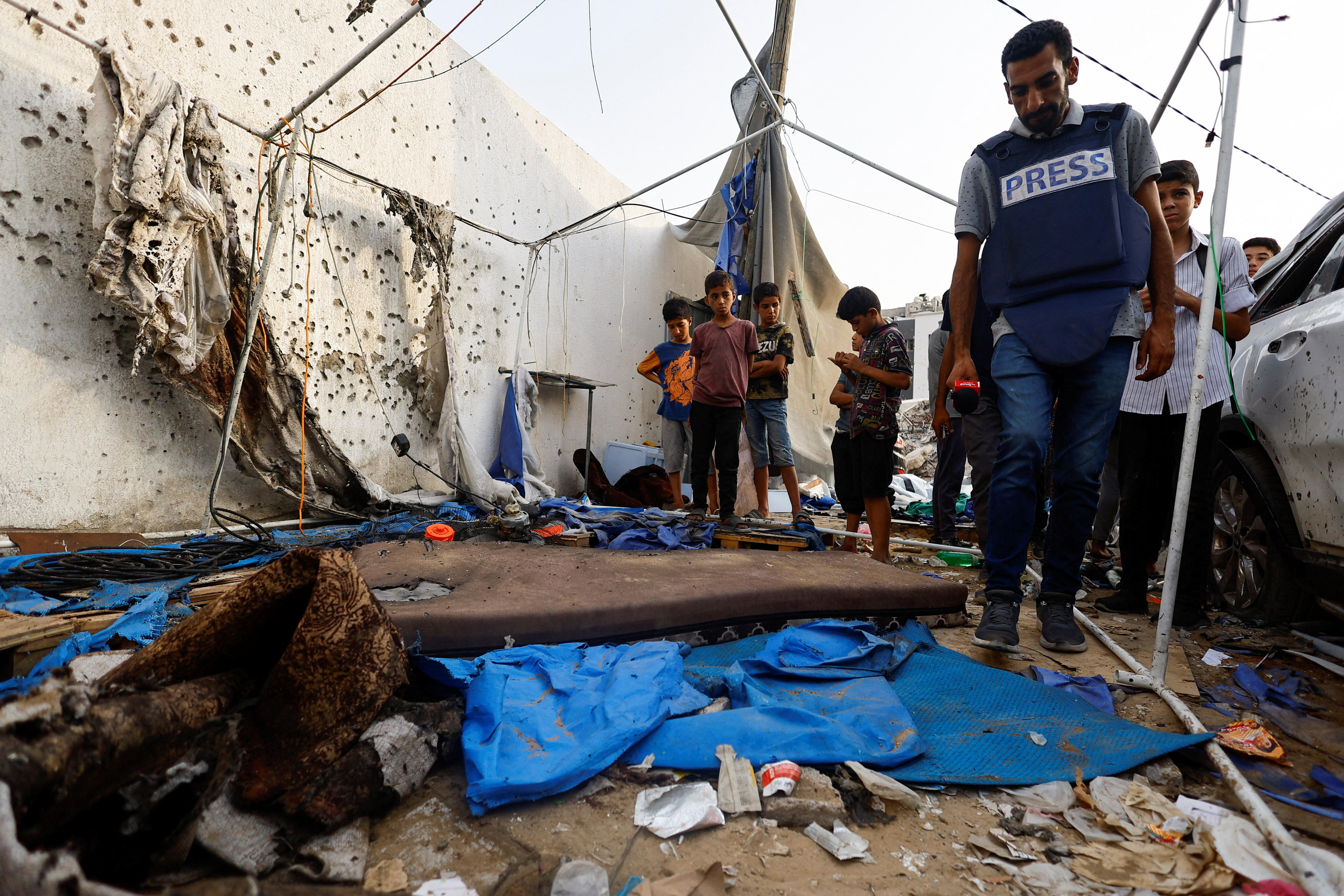 A man in a bulletproof vest marked "Press" looks through rubble inside a damaged tent frame.