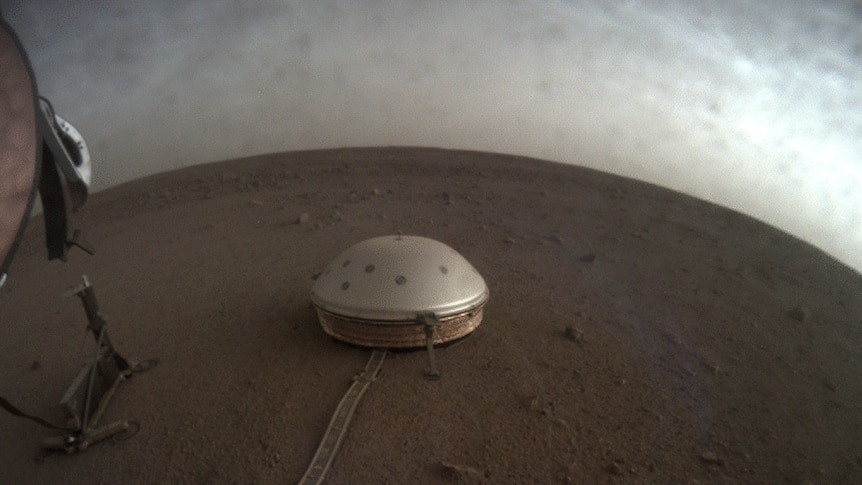 A small silver dome-shaped seismometer is seen from above on the red dirt of Mars