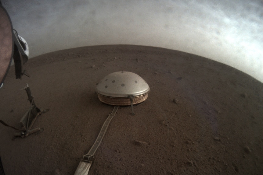 A small silver dome-shaped seismometer is seen from above on the red dirt of Mars