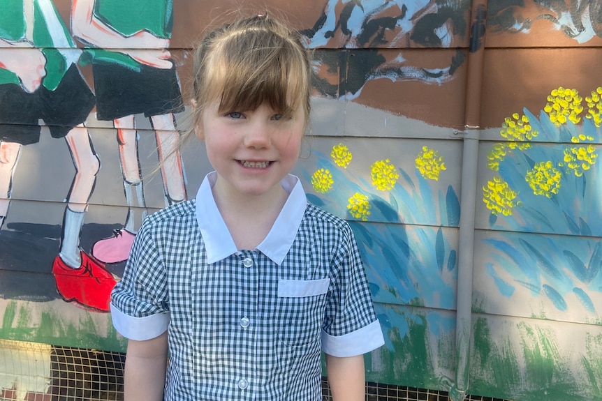 A young girl with a fringe smiles at the camera in a school uniform in front of a colourful mural.