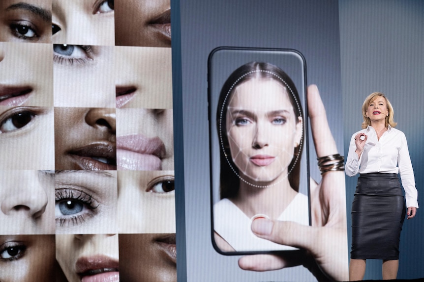 A wide shot of a woman standing on a stage giving a keynote speech. Behind her are images of faces and a hand holding a phone