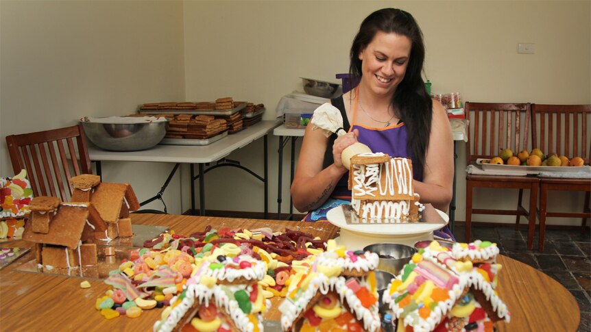 Lady with long brown hair ices a gingerbread house at a kitchen table.