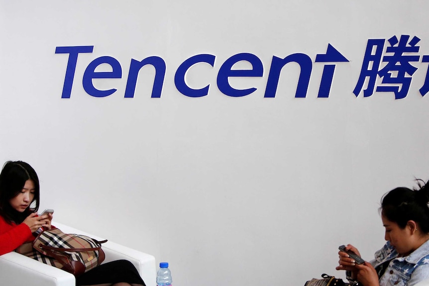 Two women use their smartphones while sitting underneath the logo of Tencent.