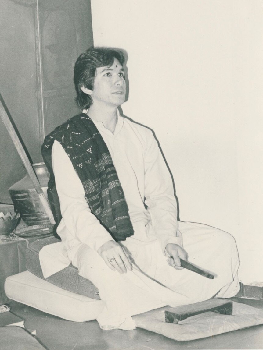 Black and white image of a man in traditional kurta and scarf, with legs crossed.