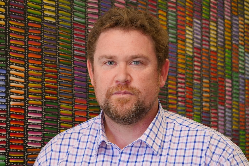 A man with a beard stands in front of a colourful painting