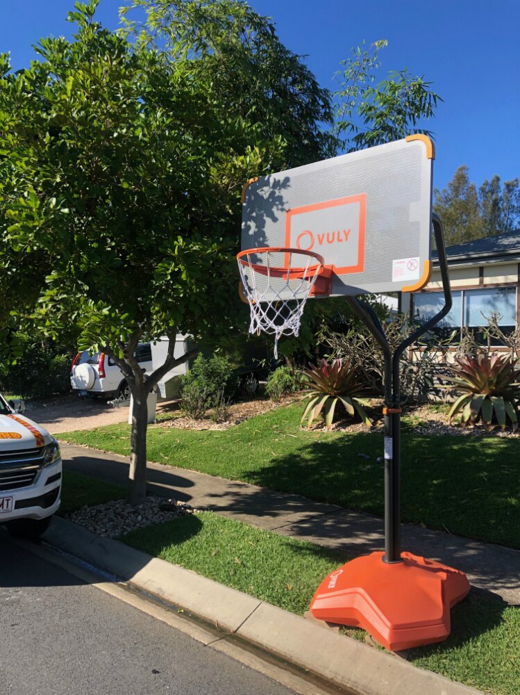 A temporary basketball hoop sits between a footpath and the road in front of a house.