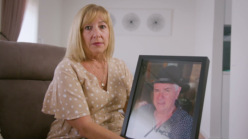 A woman looking sad at the camera while holding a photo of an older man