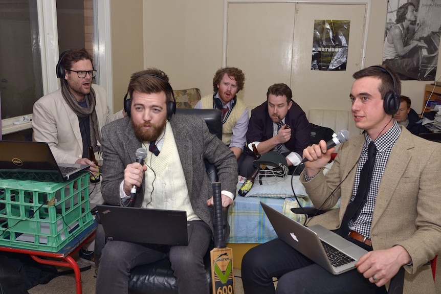 Wide shot of a group of men broadcasting in a lounge room.