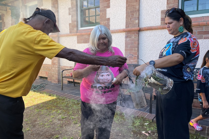An Indigenous woman surrounded by two people holding cans and smoke circling the air