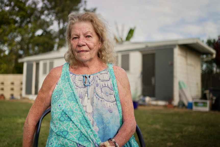 A woman stands outside a small demountable home, looking serious.