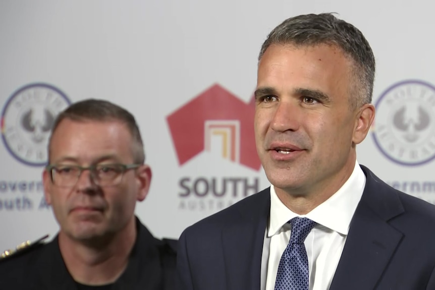 A man wearing a suit and tie with another behind wearing a blue shirt