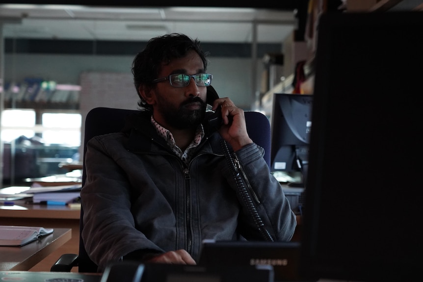 A man wearing glasses sits at a desk on the phone and looking at a desktop computer