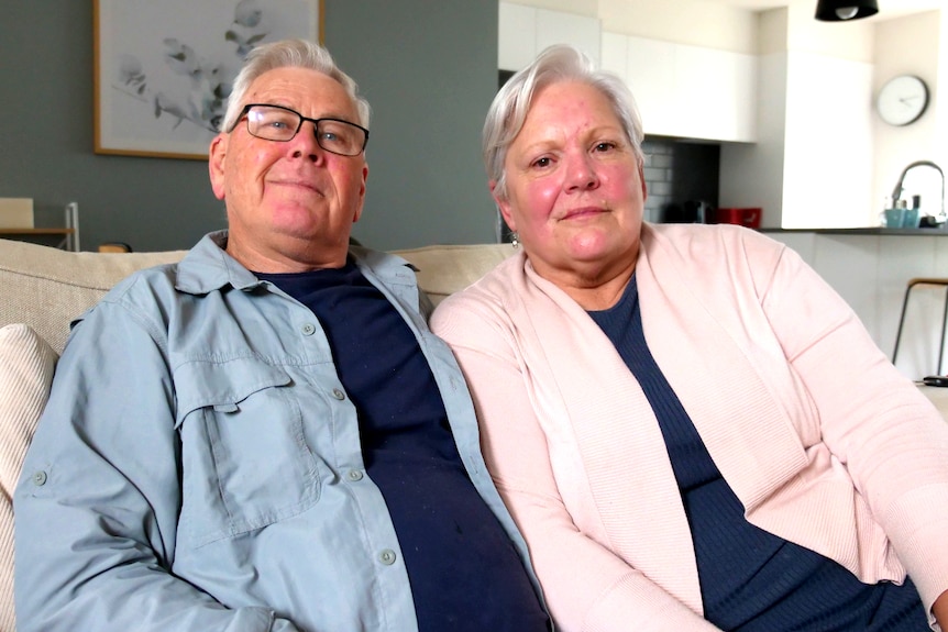 A man and a woman, grey hair slightly smile, sit close on the couch. Painting on grey wall behind, kitchen bench to the side.