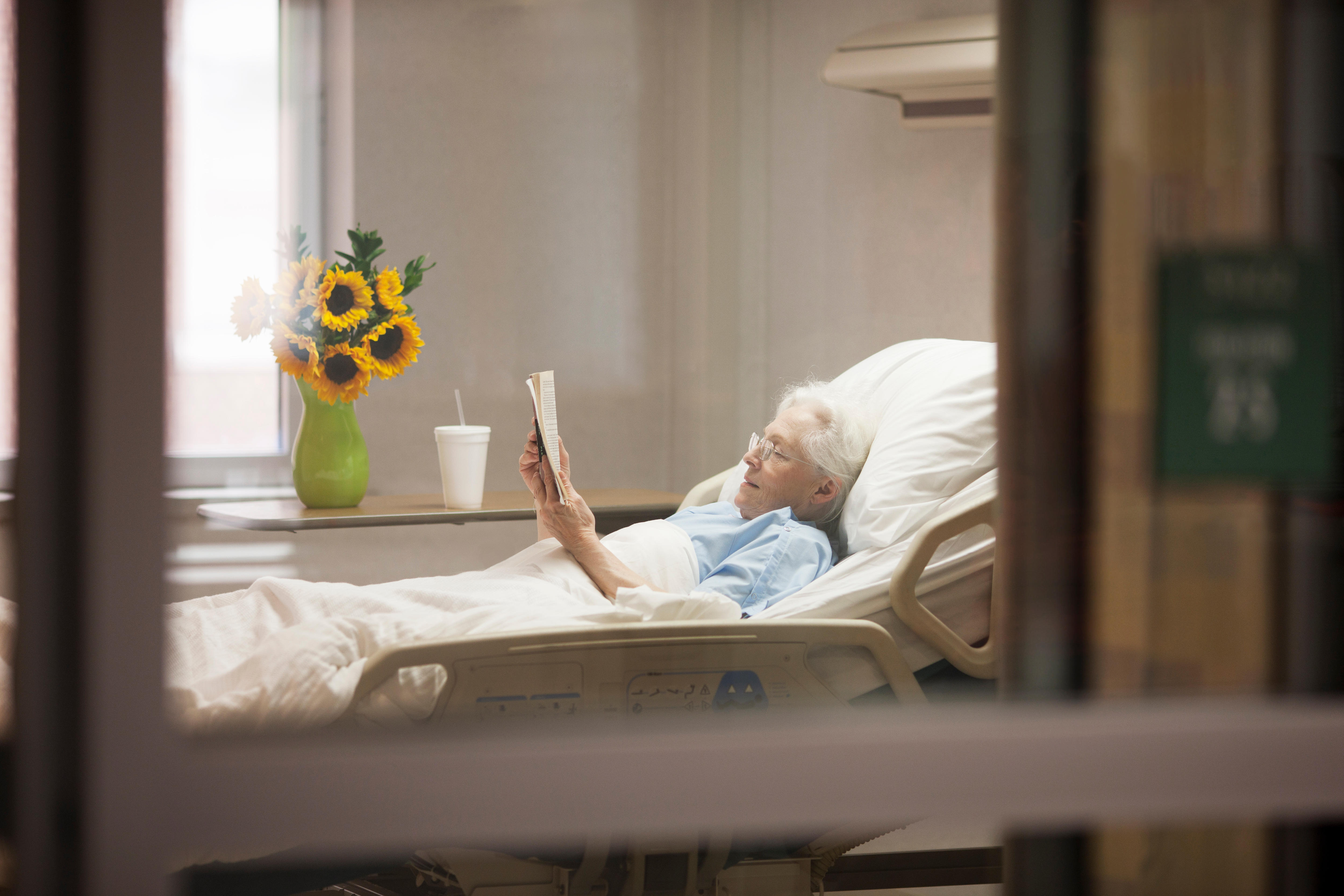 70s woman with glasses reading a book while lying in a hospital bed in her room