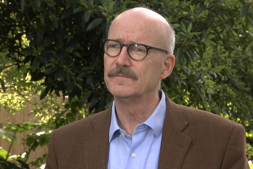 A man in a brown jacket standing in front of a hedge