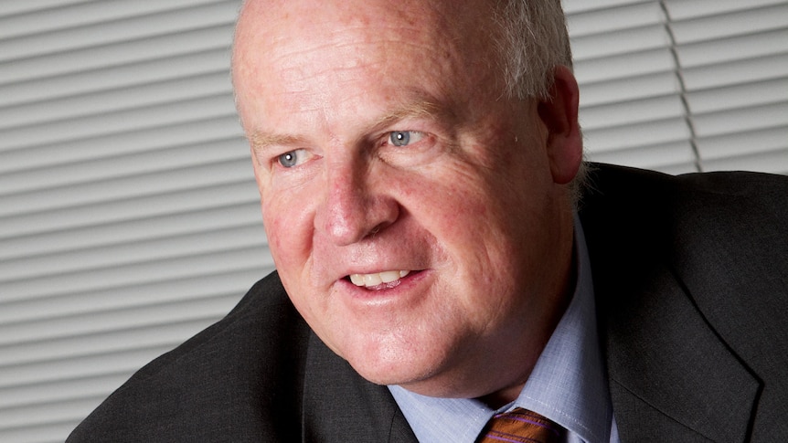 A bald-headed man in a suit smiles, looking away from the camera.