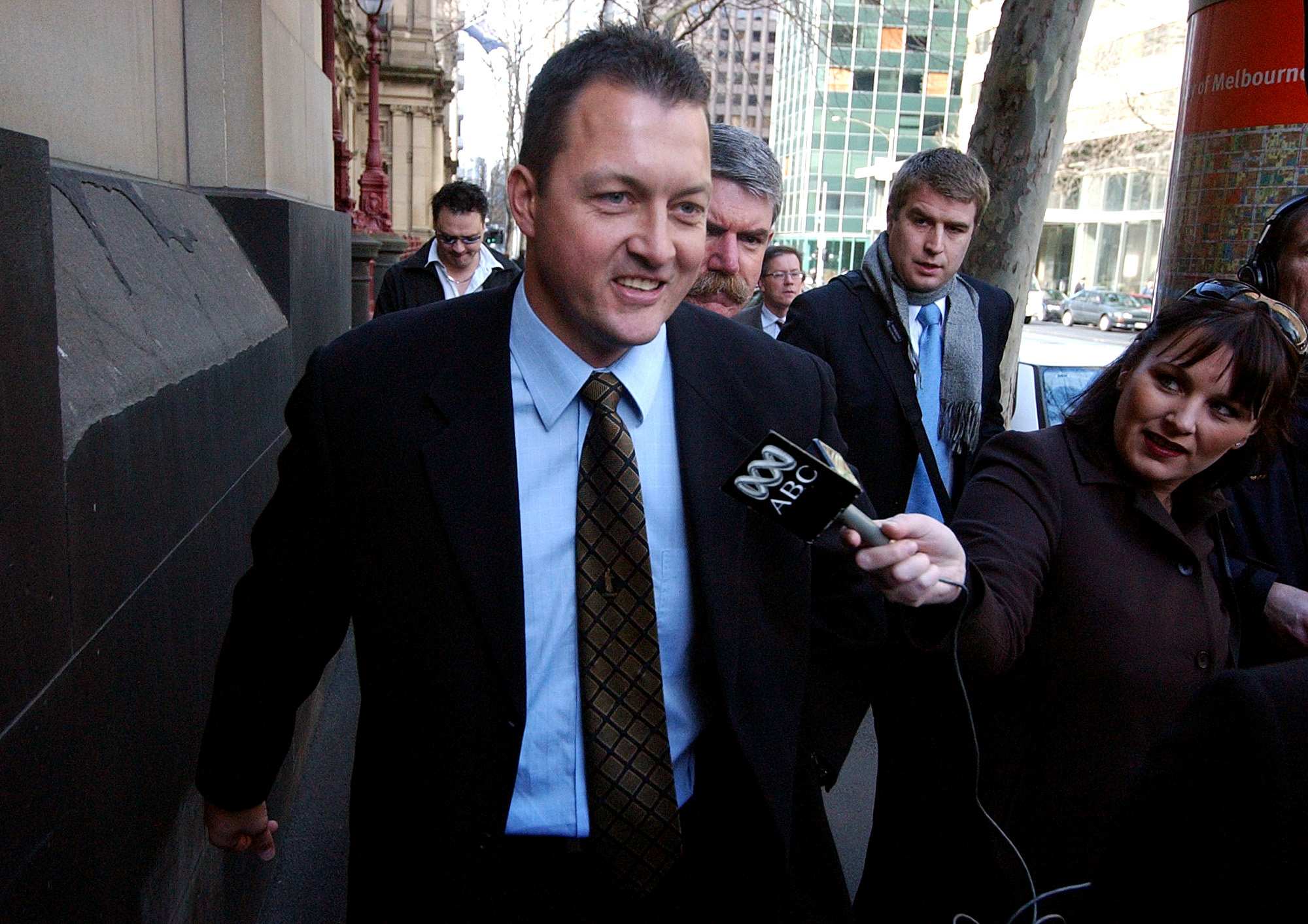 Paul Dale walking down the street in a suit, surrounded by journalists.