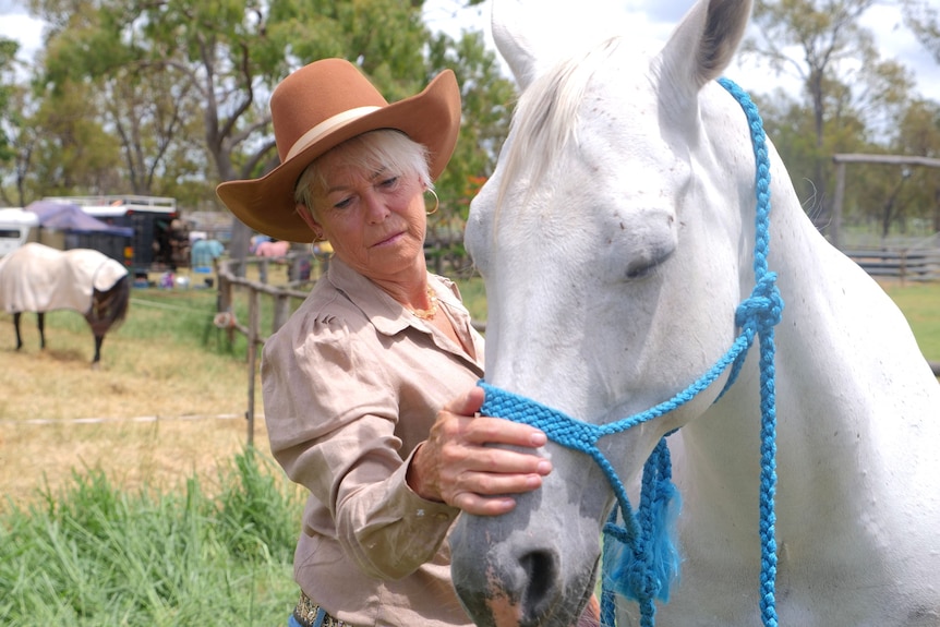 A woman with short blonde hair wearing a big brown hat stands beside a white horse and strokes its nose.