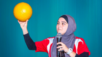 Soccer player Natasha Hill holds a soccer ball, she is wearing headscarf and holding a mic to her face.