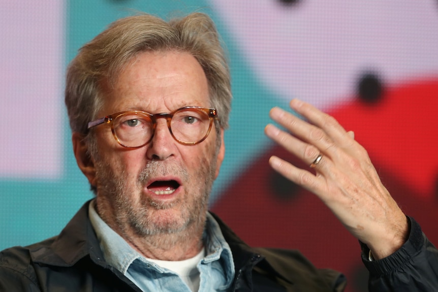 A man in tortoiseshell glasses speaks while lifting his hand.