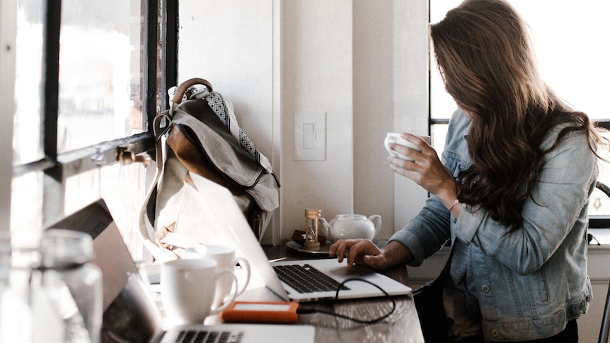 A woman sits at a computer with her hair shielding her face, drinking a coffee.