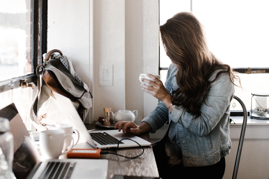 A woman sits at a computer with her hair shielding her face, drinking a coffee.