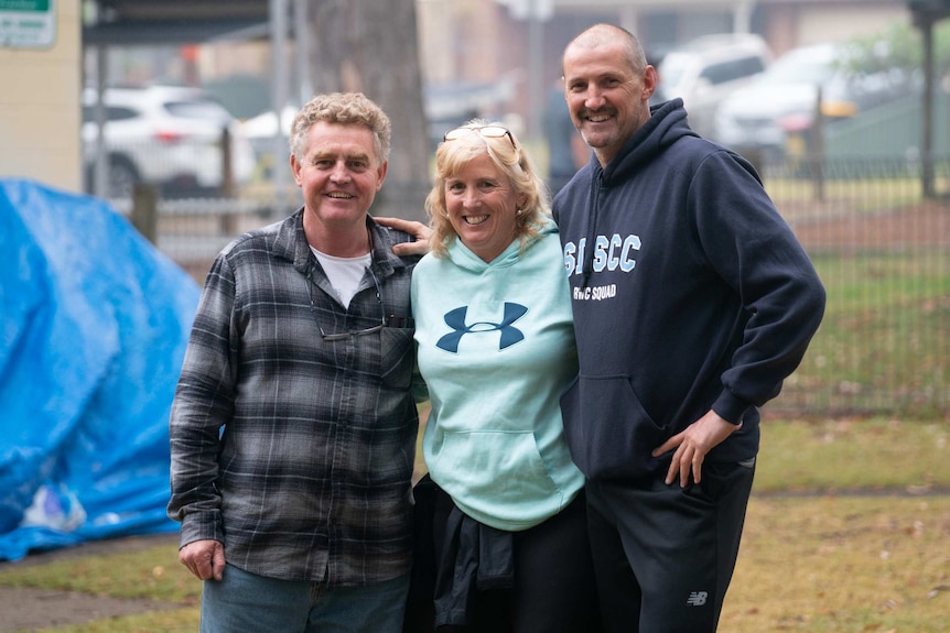 Two men and a woman stand with arms around each other in the grounds of a camping ground.