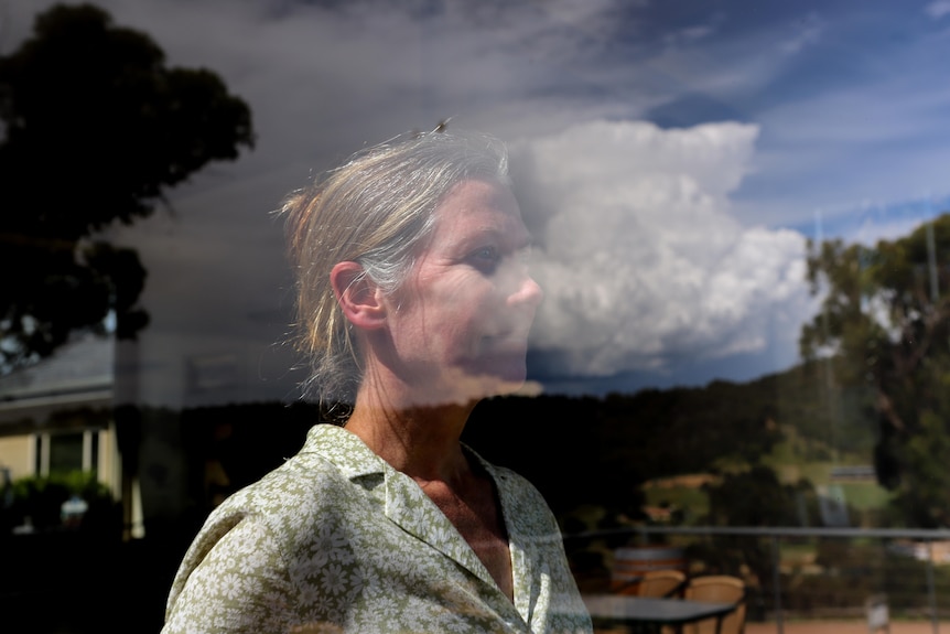 Woman with fair hair wearing cream blouse stares out window with reflection of hills in distance