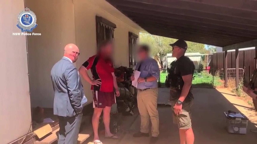 Four men, three police and one civilian, in a group under an awning outside a home.