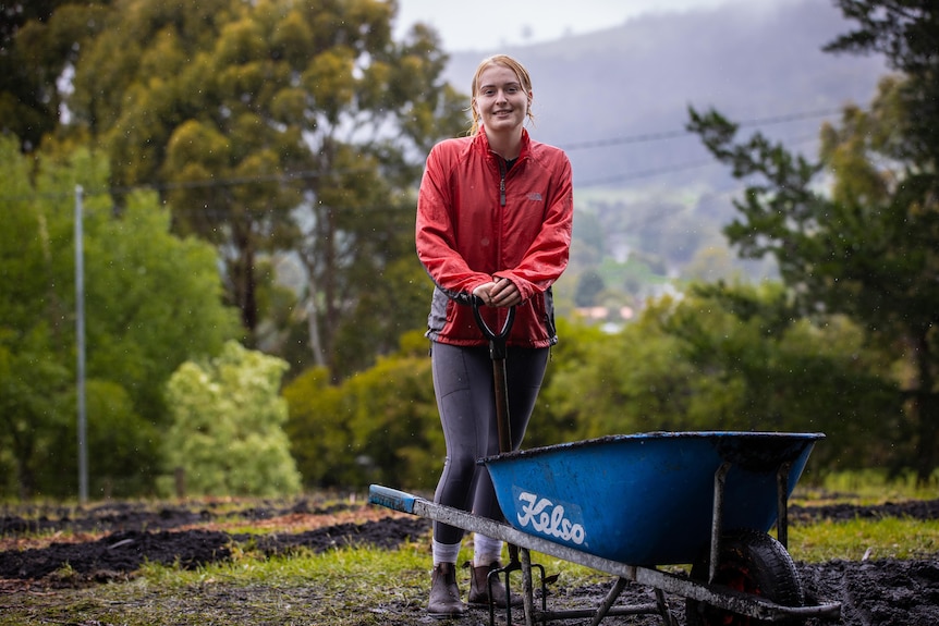 A woman wearing a red jacket stands behind a wheelbarrow, smiling.