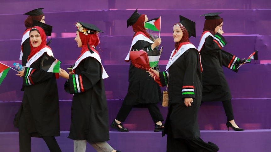 Six women graduates wearing mortar boards and academic gowns carrying Palestinian flags cross at stage at a graduation ceremony