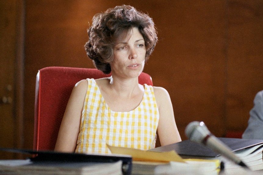A woman with short hair and a yellow gingham dress sits behind a court table. 