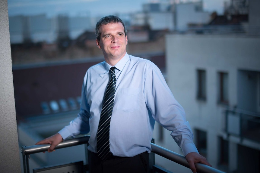 A man in blue collared shirt and tie leans against modern balcony railings and looks out across a dark city.