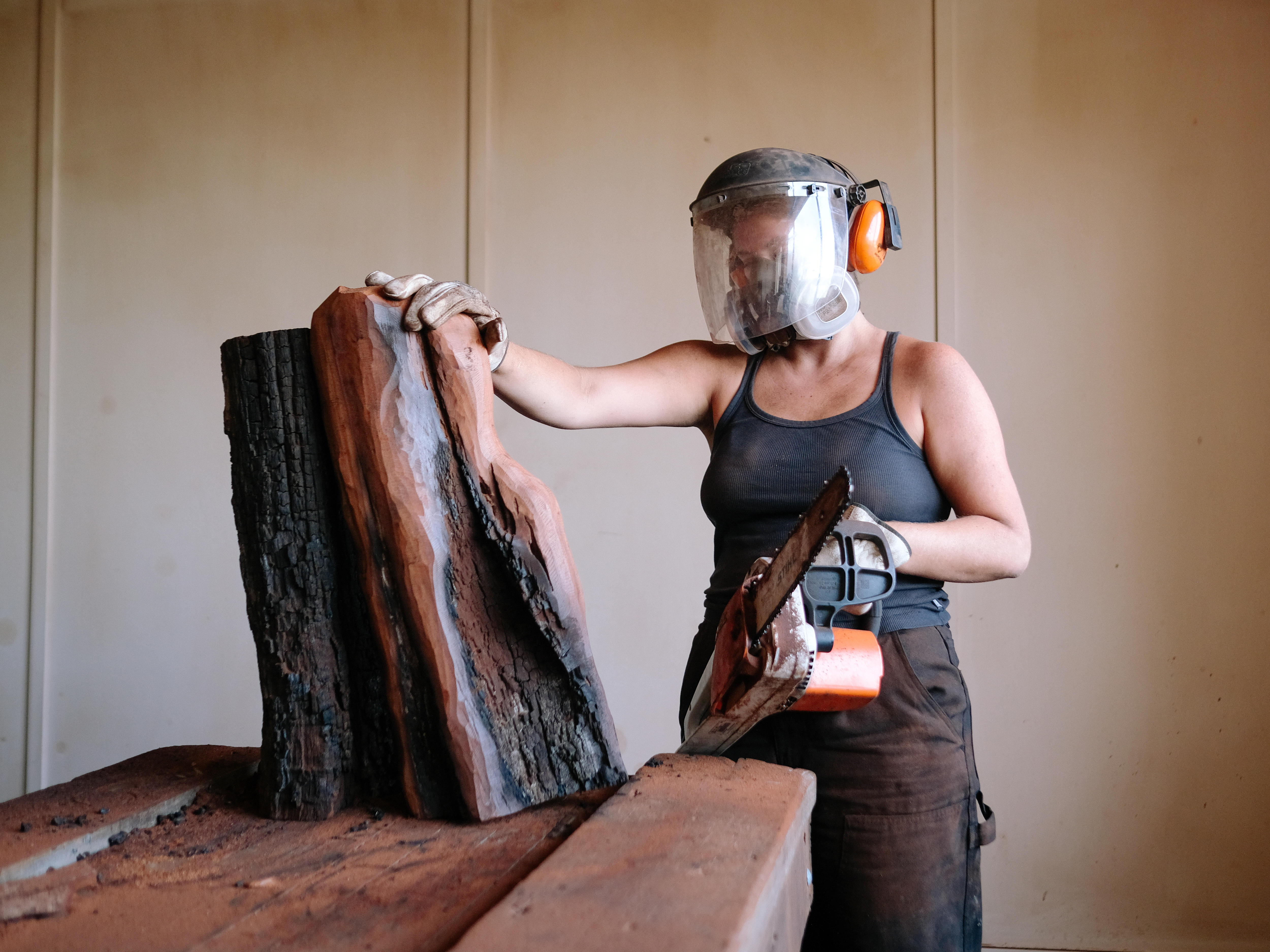 Artist in process of making a sculpture from wood, holding a chainsaw and wearing a protective face mask and earmuffs