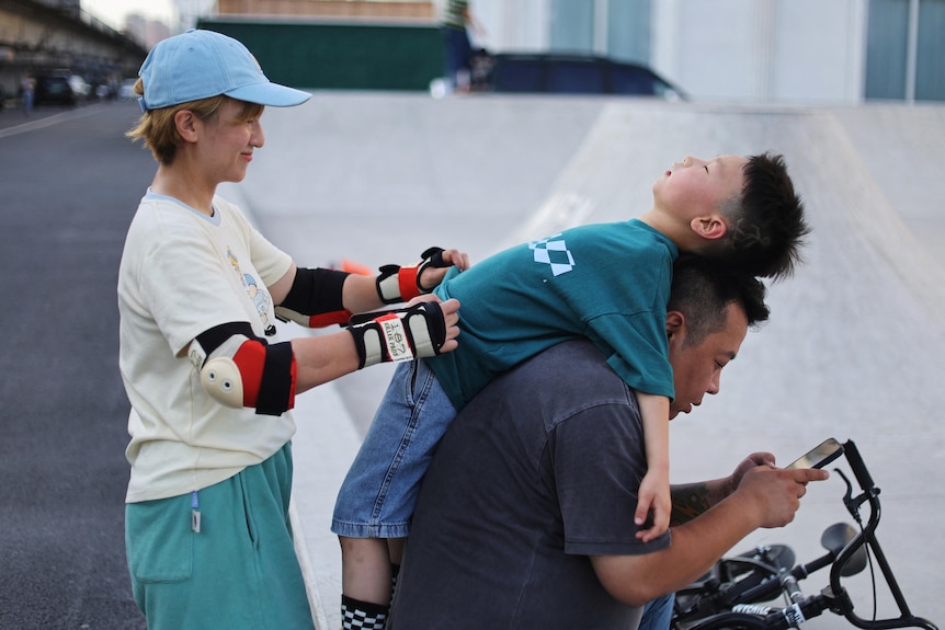 A woman with elbow pats on tends to her young child, a boy, who is laying on his dad's back as his dad sits on a bike. 
