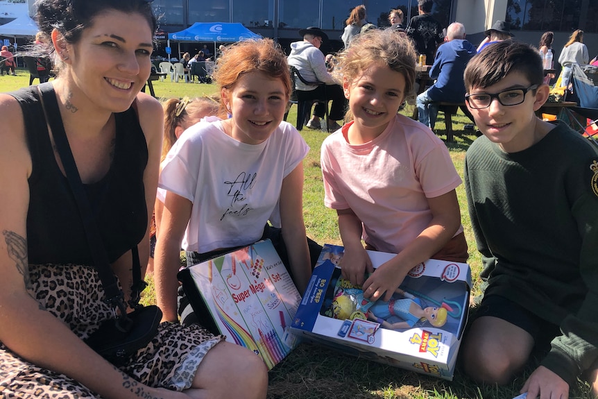 A woman sits with two girls and a boy on a lawn. The girls are holding toys.