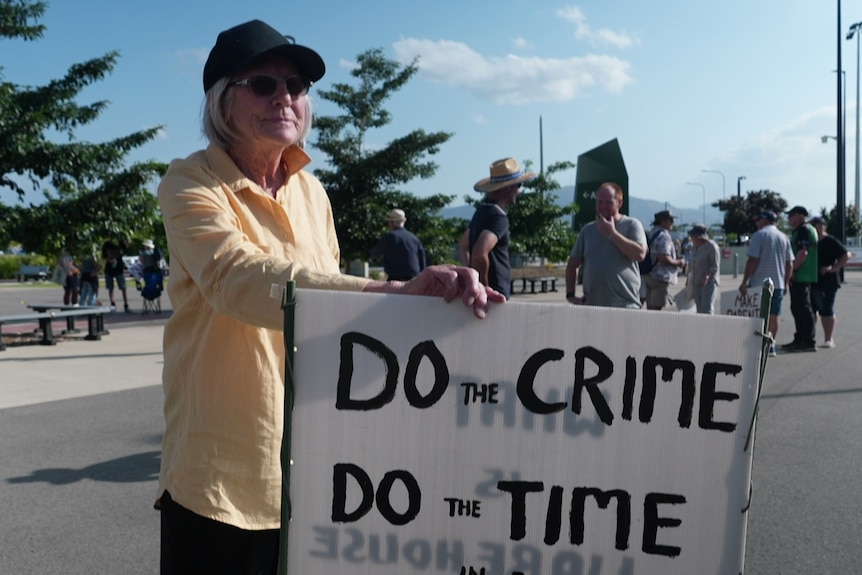 A woman in a long sleeve shirt cap and sunglasses holds a sign. 