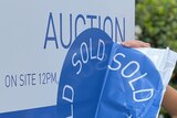 A smiling brown-haired woman puts a 'sold' sticker on a 'for auction' sign outside a house.