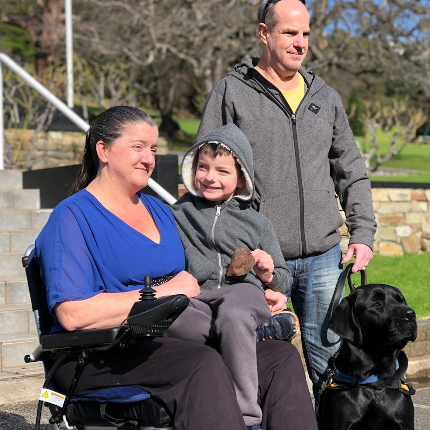 A woman in a motorised chair holds a young boy on her lap. A man stands behind them holding a dog on a lead