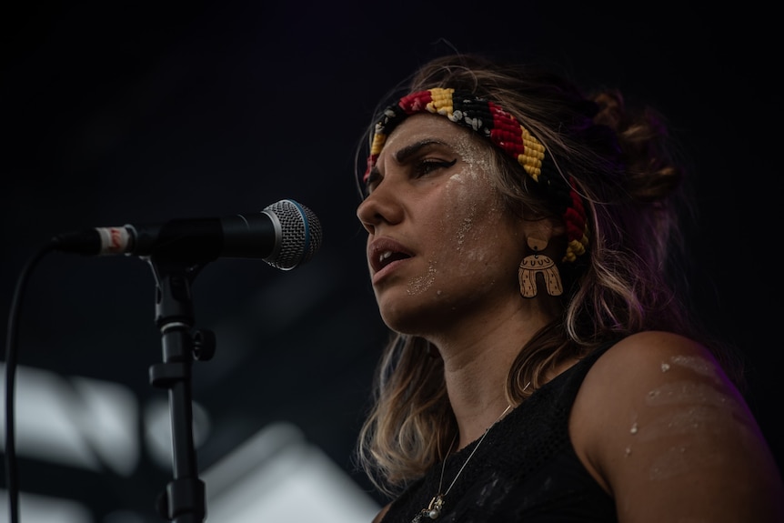 Kirli Saunders stands at a microphone while wearing a headband in the colours of the Aboriginal fla