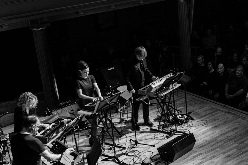 A black-and-white photo of members of the Will Gregory Moog Ensemble playing in a concert-hall in front of an audience;