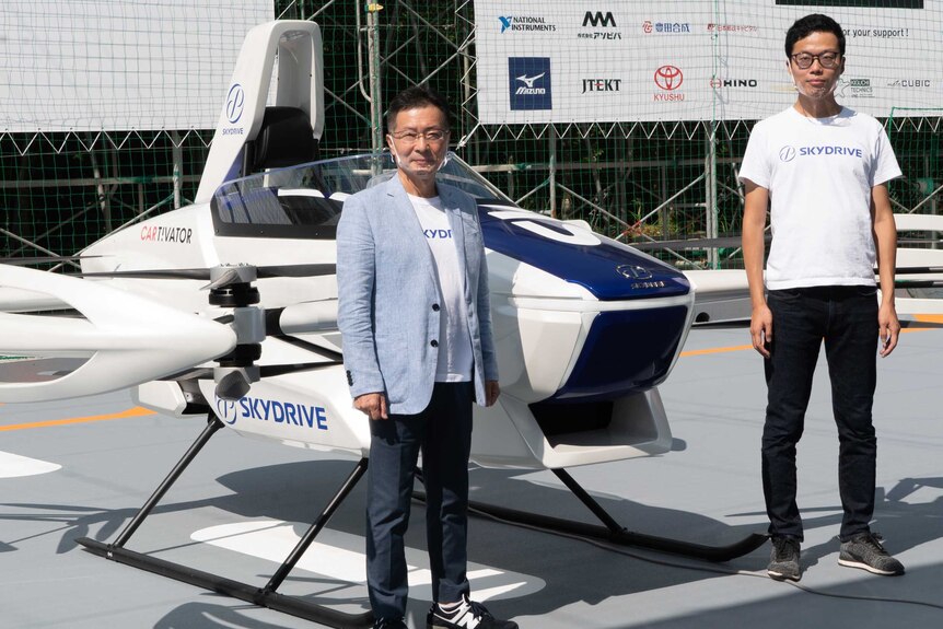 Two men standing next to a flying car prototype
