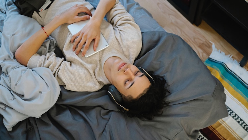 A young person is laying on top of a bed with over-ear headphones on and a tablet laying on their chest.