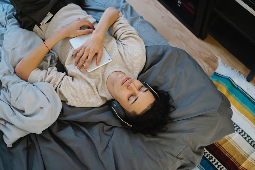 A young person is laying on top of a bed with over-ear headphones on and a tablet laying on their chest.