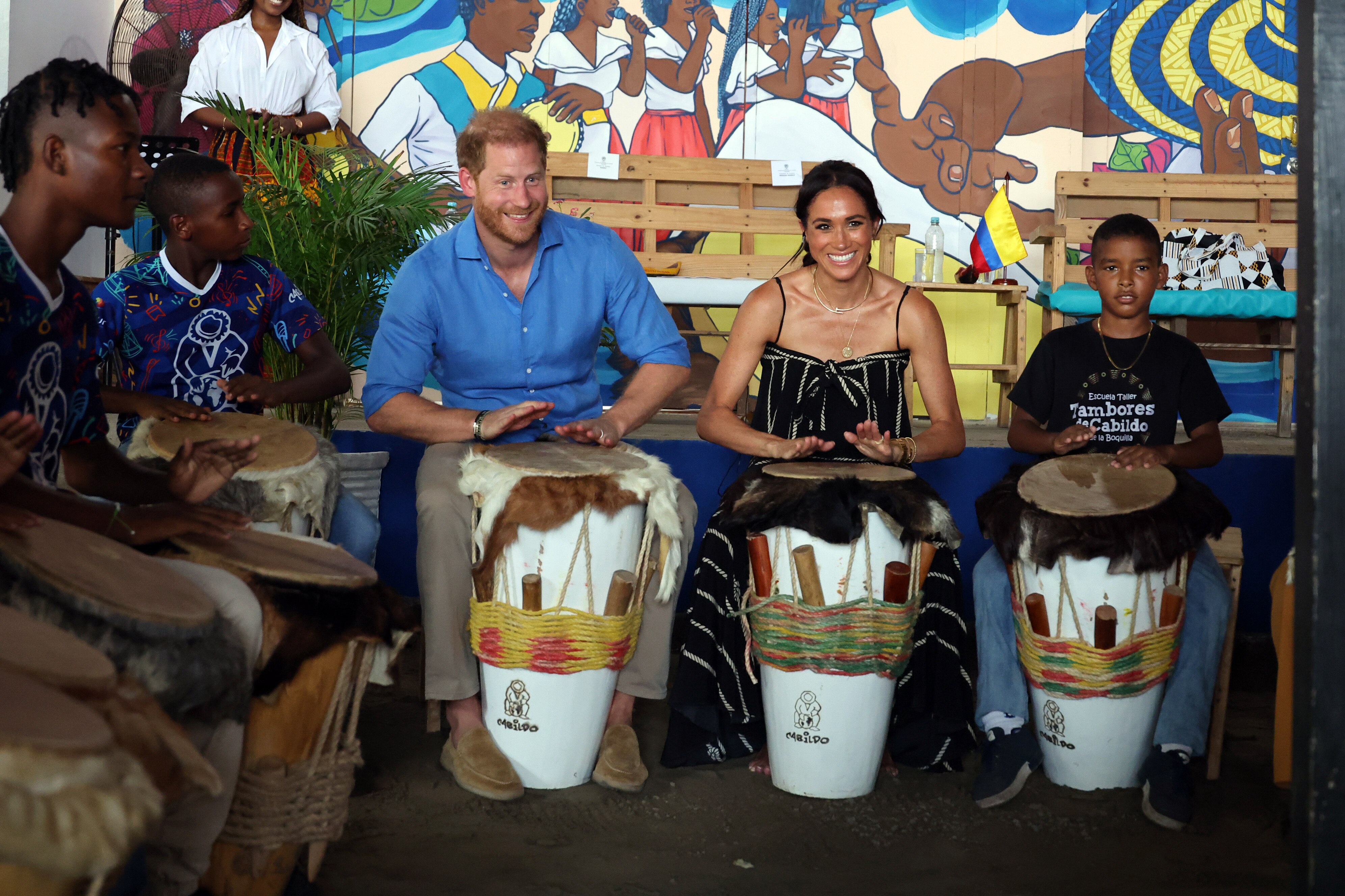 Harry and Meghan smiling broadly as they take part in a group drumming class.